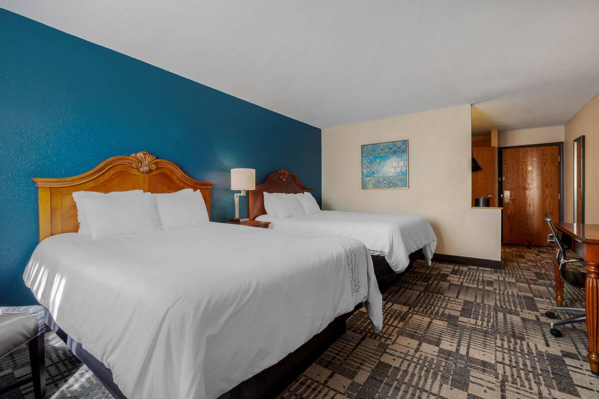 A hotel room with two beds, a desk, a chair, and a painting on the wall.