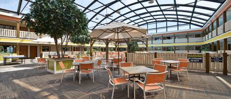 A spacious indoor area with a high ceiling and a glass roof, featuring multiple dining tables and chairs, a central tree, and a warning sign about no lifeguard on duty.