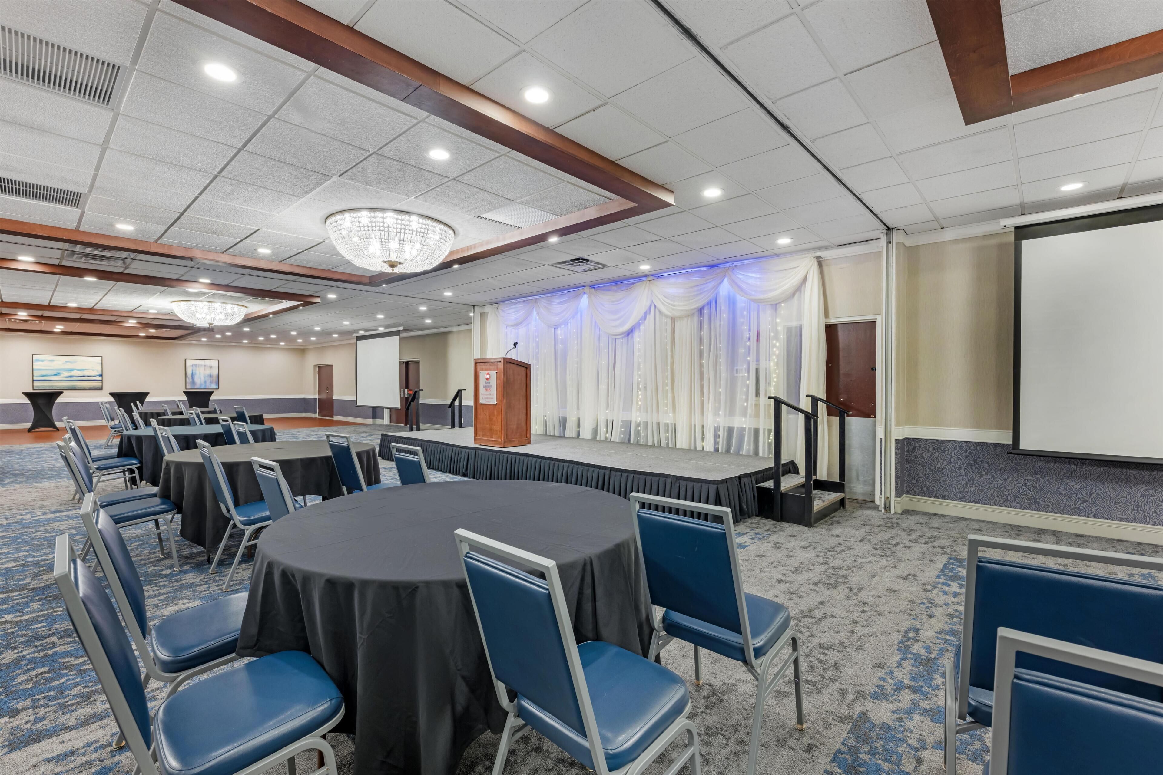 A conference room with round tables, blue chairs, a podium, and a large screen.