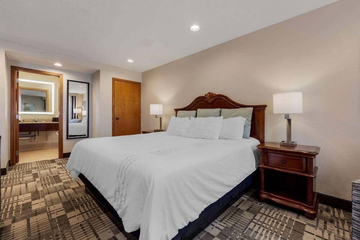 A hotel room with a large bed, bedside lamps, a wooden nightstand, a mirror, and a bathroom visible through an open door.