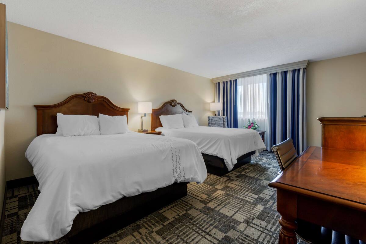 A hotel room with two beds, a wooden headboard, a small table, and a view of the city through large windows.
