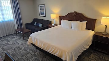 Una habitación de hotel con una cama grande, dos mesitas de noche con lámparas, un sillón oscuro, una mesita pequeña y una alfombra estampada.