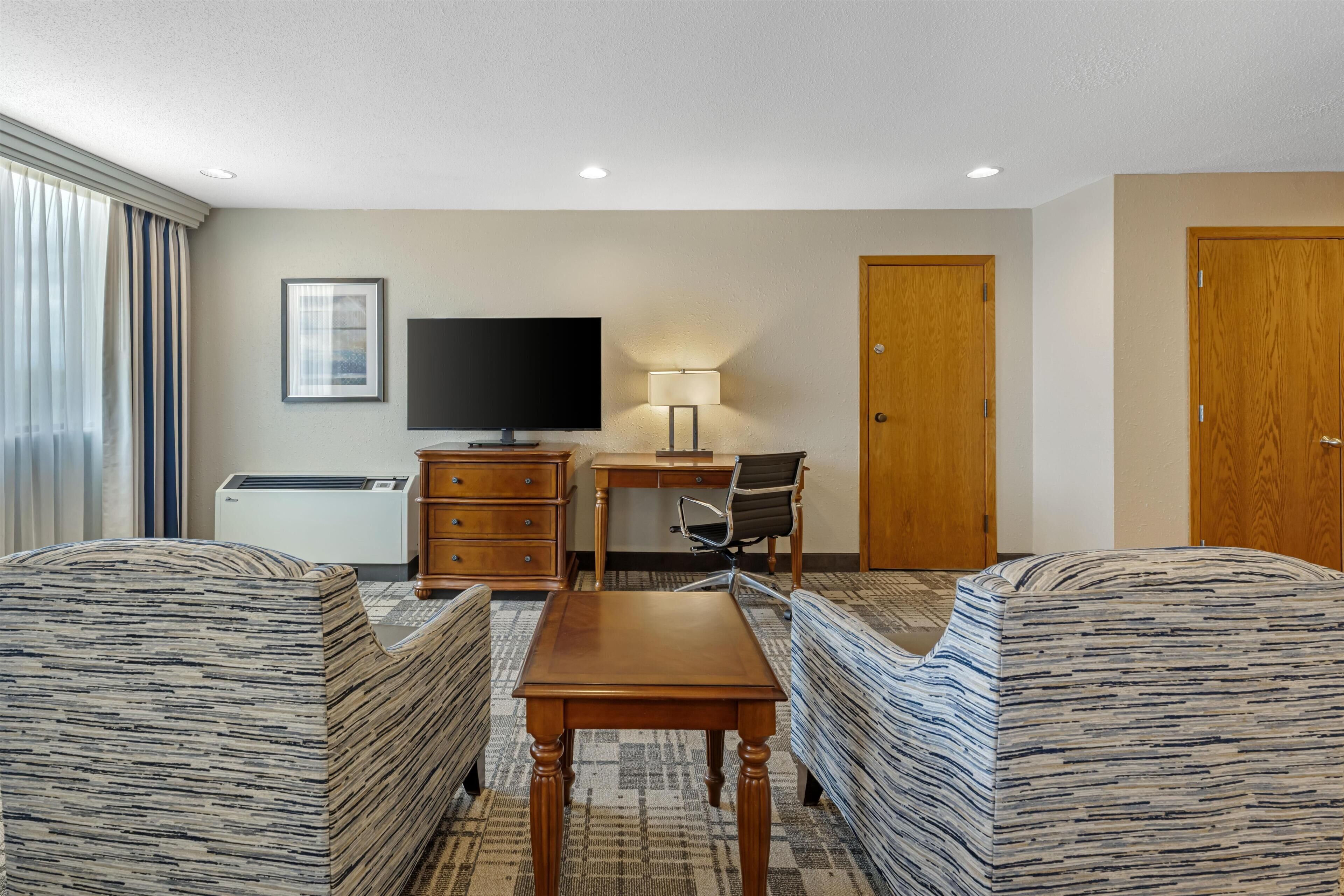 A hotel room with a bed, a desk with a chair, a television, a lamp, and a wardrobe.