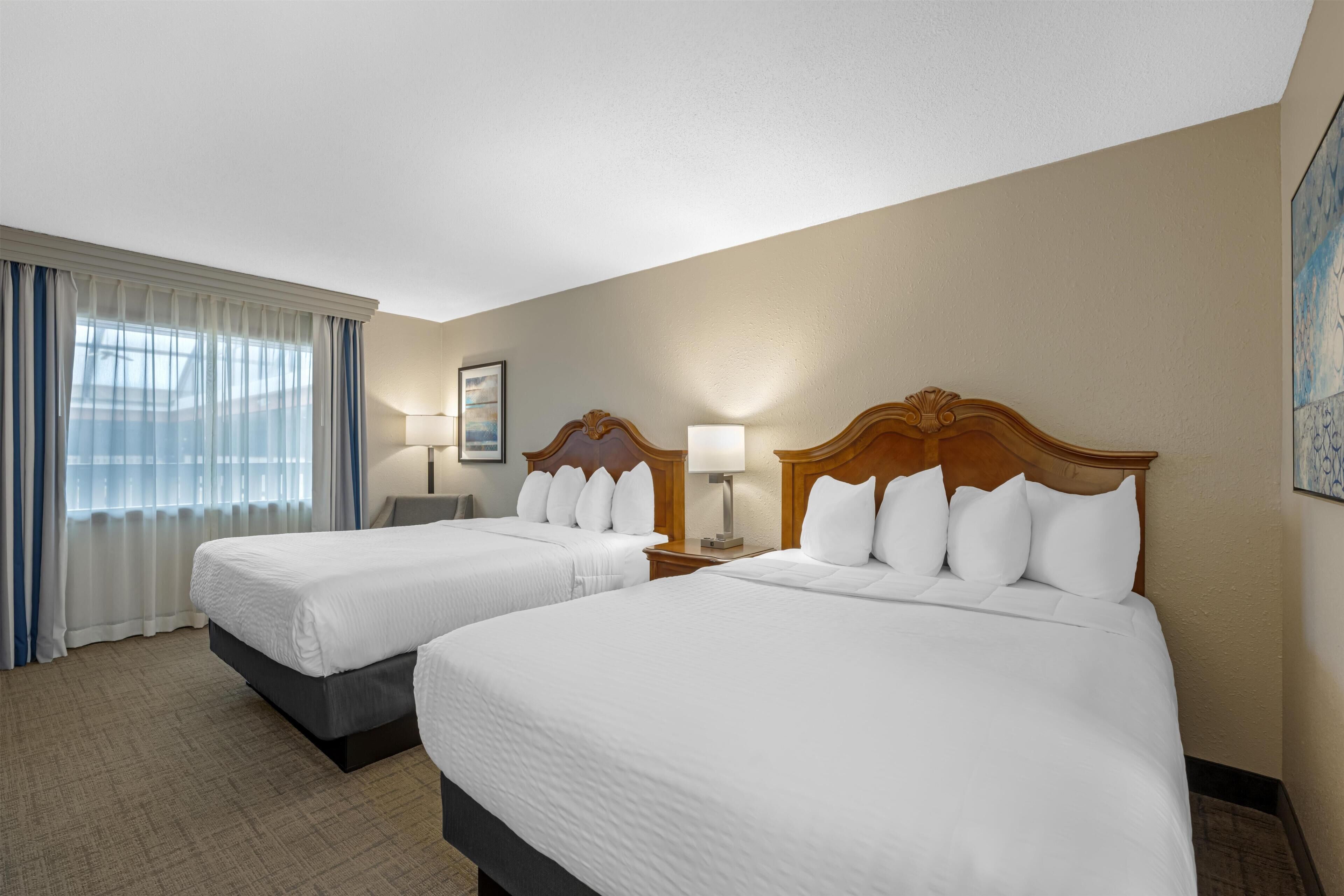 Habitación de hotel con dos camas, un ventanal con cortinas, un cuadro en la pared y una lámpara en cada mesita de noche.