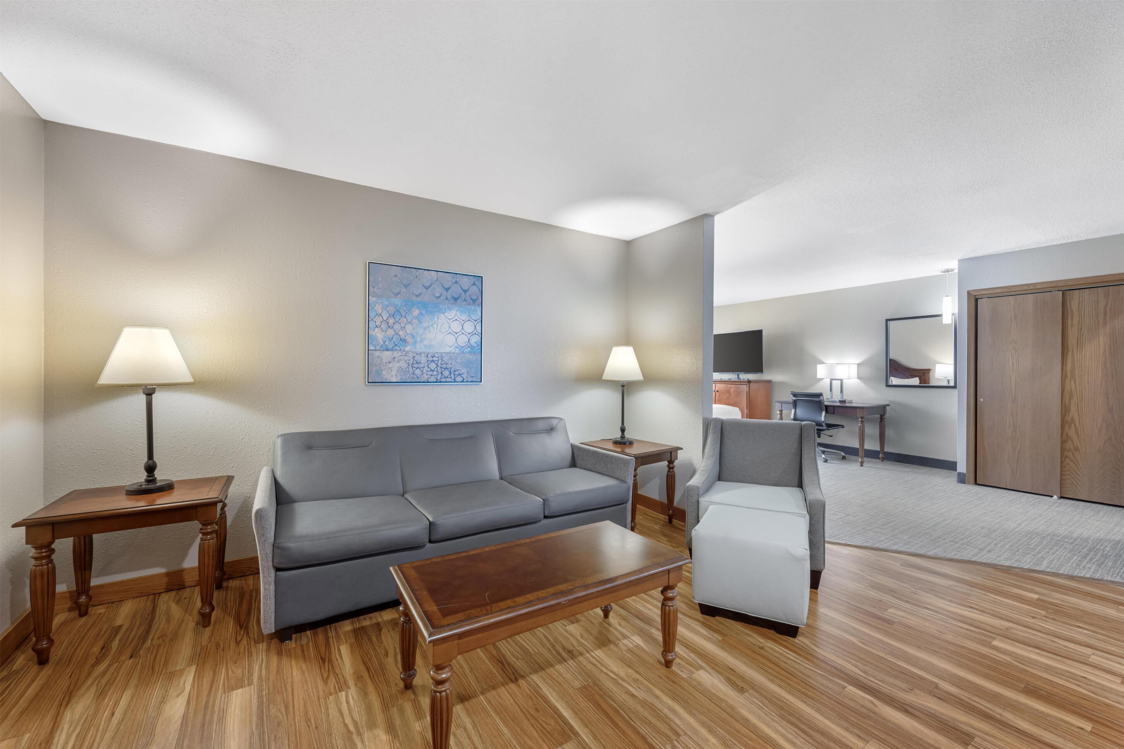 Ein modernes Hotelzimmer mit grauem Sofa, einem Holztischchen, zwei Lampen, einem Gemälde, einem Schreibtisch und einem Kleiderschrank.