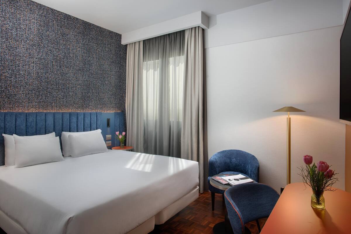 A hotel room with a large bed, a chair, a small table, a lamp, and a vase with flowers.