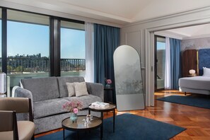 A modern hotel room with a grey sofa, a round coffee table with a vase, a large mirror, and a view of the city through large windows.