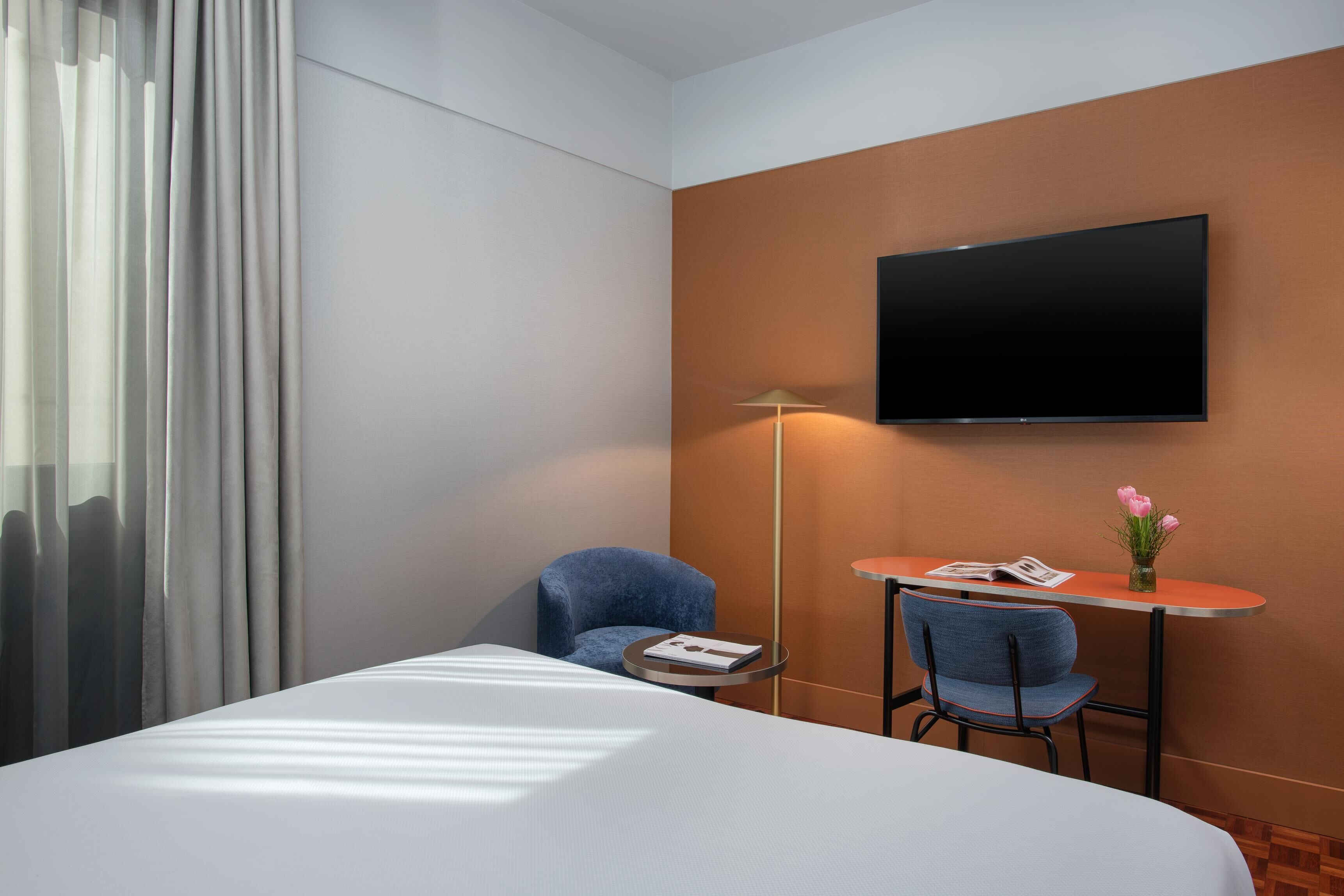 Una camera d'albergo con un letto, una TV a parete, una scrivania con una sedia e una lampada.
