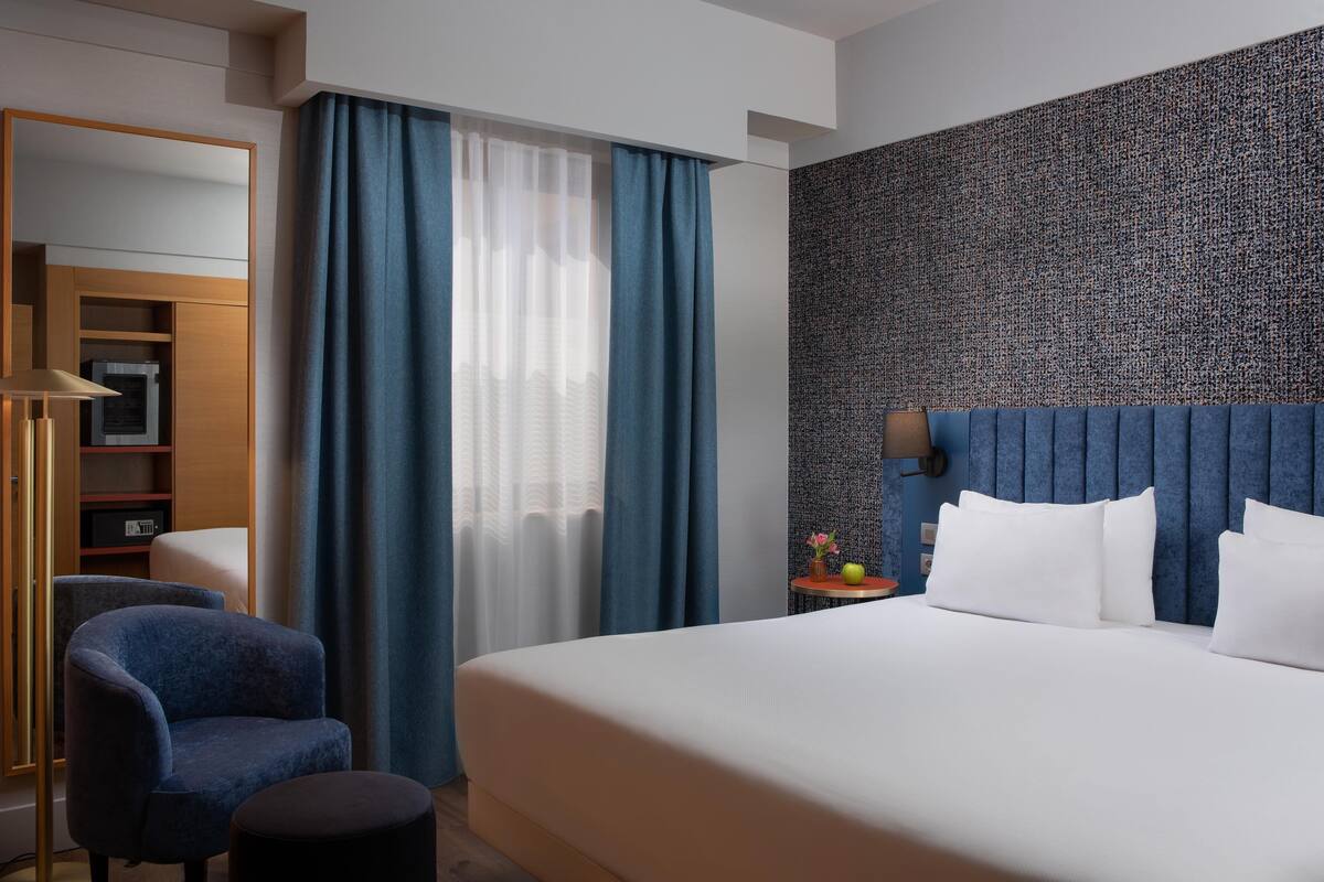 A hotel room with a large bed, a blue upholstered chair, a small round table with a vase, and a wall with a textured pattern.
