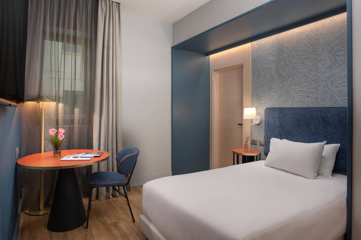 A hotel room with a bed, a round table with a chair, a small table with a lamp, and a window with curtains.