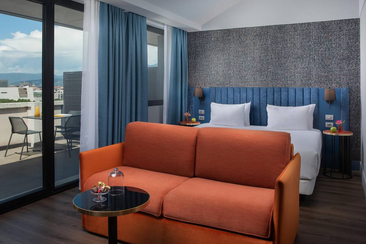 A modern hotel room with a large bed, orange sofa, and a balcony with a table and chairs.