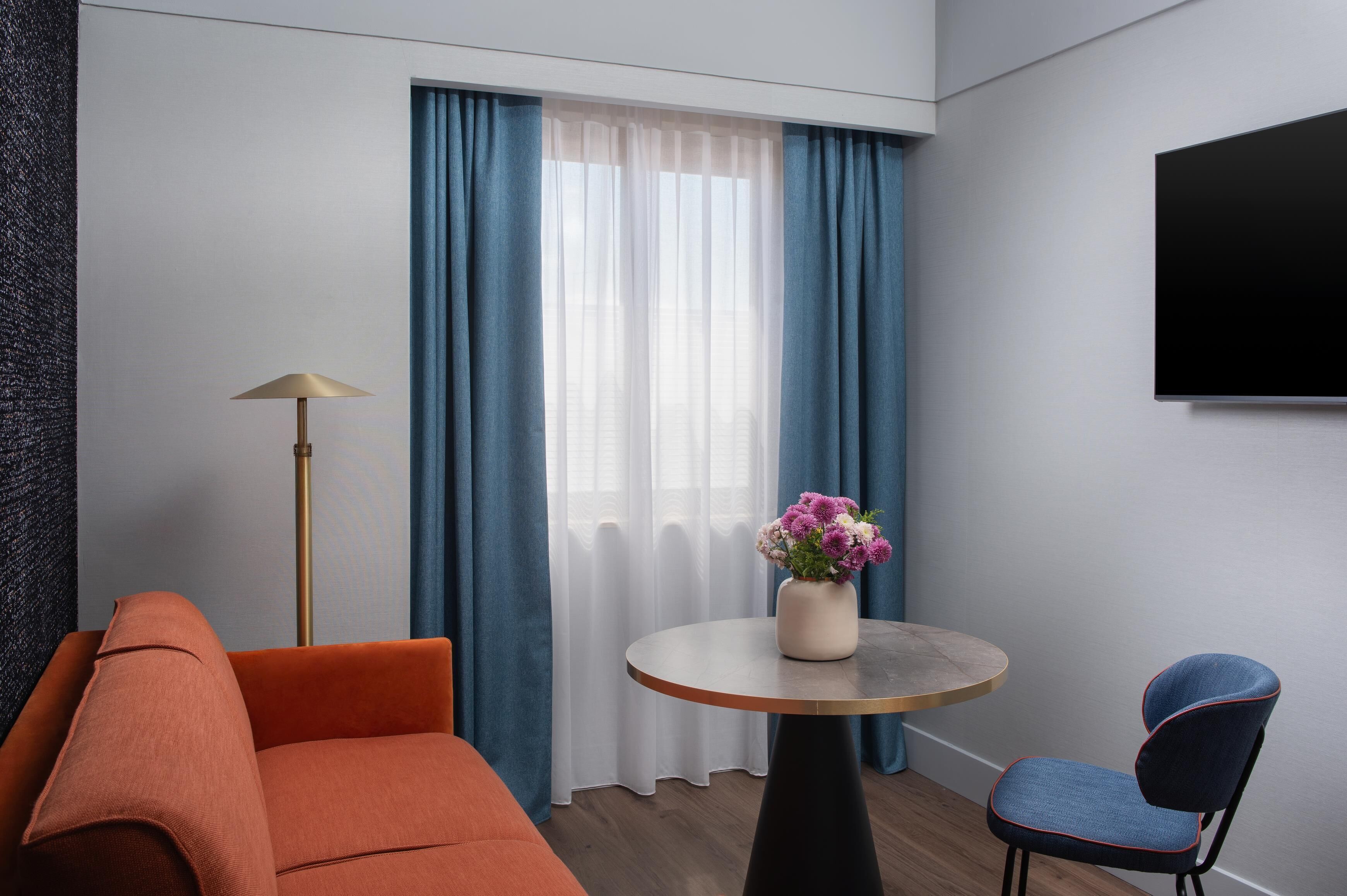 Une pièce moderne comprenant un canapé, un fauteuil, une petite table ronde ornée d’un vase de fleurs, un lampadaire et un téléviseur fixé au mur.