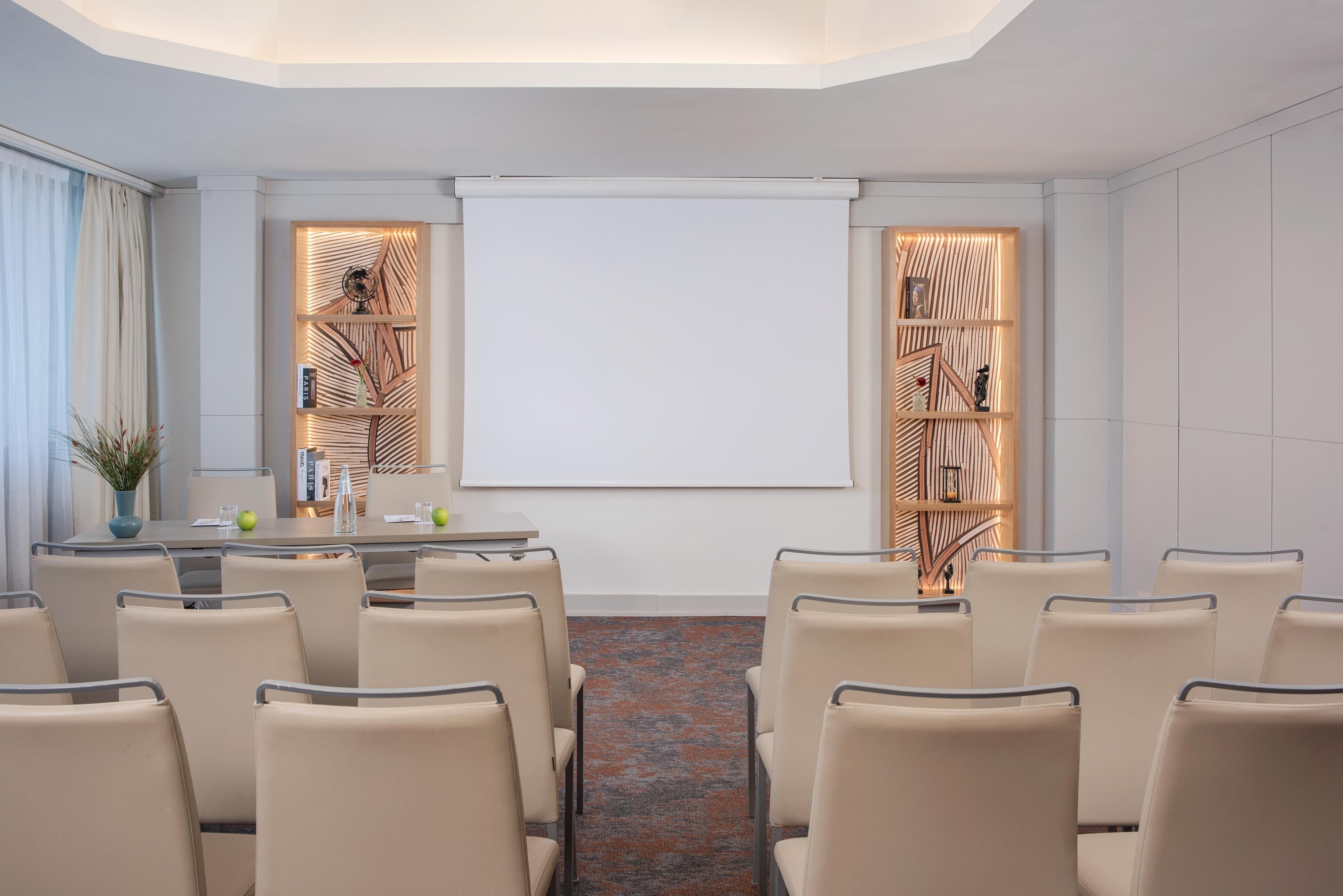 A conference room with a whiteboard, beige chairs, a table with a vase and apples, and wooden shelving with decorative items.