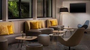 Un espacio lounge moderno con sofás, otomanas y mesas redondas, un televisor de pantalla plana y un ventanal con vistas a la vegetación.