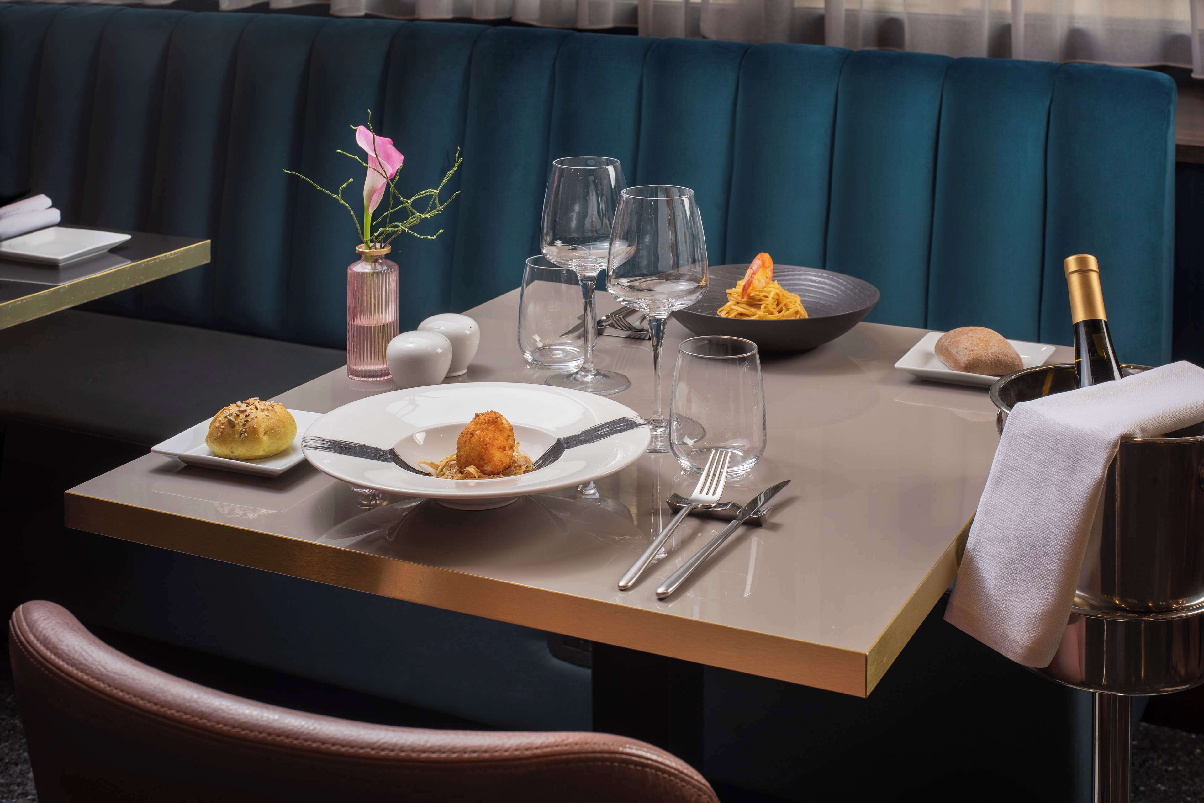 A dining table set with plates, cutlery, glasses, and a bottle of champagne, with a vase of flowers and a bowl of pasta on the table.