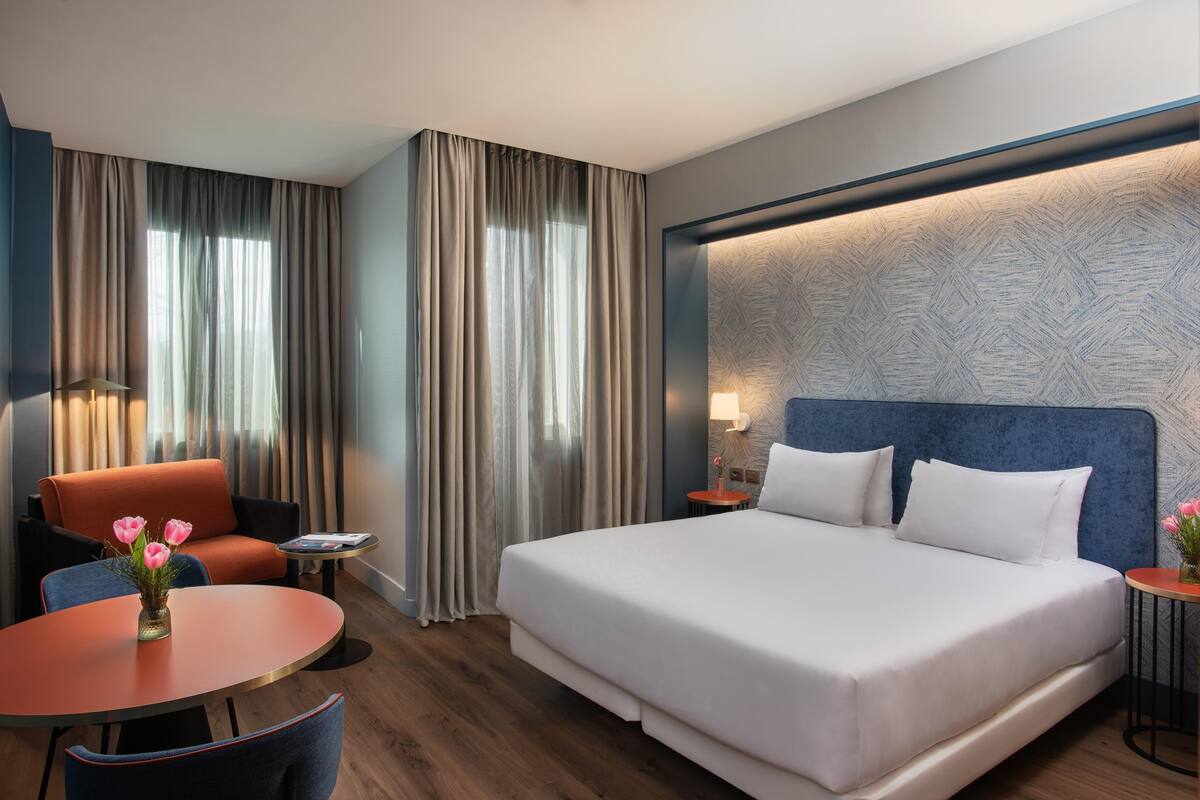 A hotel room with a large bed, a sofa, a round table with a vase of flowers, and a small side table with a lamp.