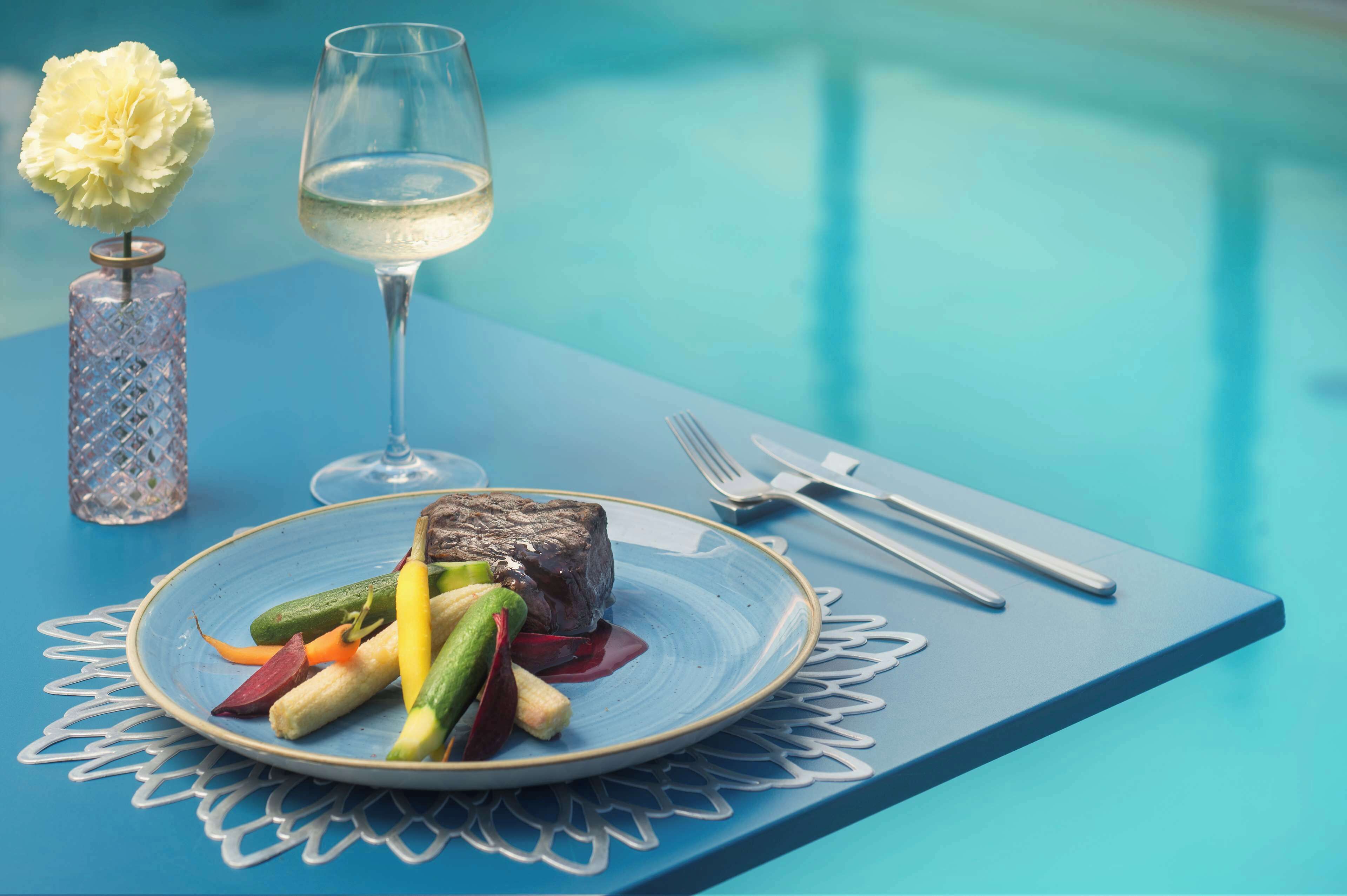 Uma mesa de jantar com um prato de comida, um copo de vinho branco e um vaso com uma flor.