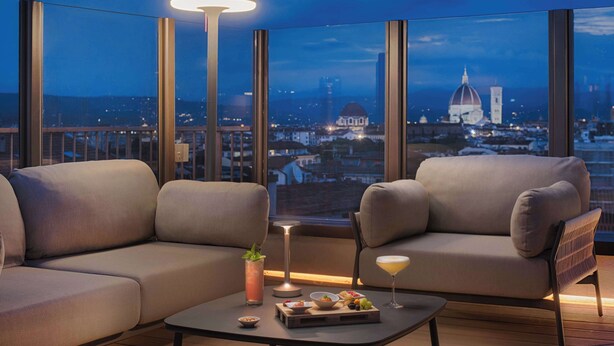 Una habitación moderna con dos sofás, una mesa de centro y una lámpara. Se aprecia un paisaje urbano con una cúpula y edificios a través de ventanas amplias.