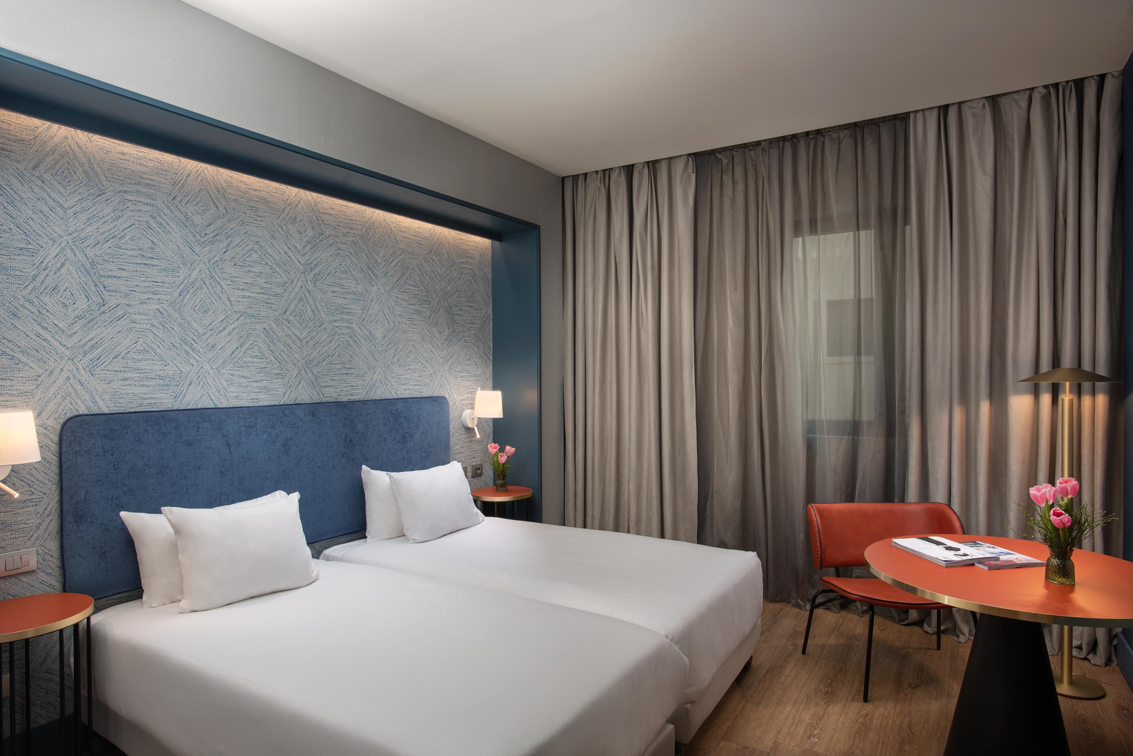 A hotel room with a large bed, a desk with a chair, a small table with a vase of flowers, and a window with curtains.