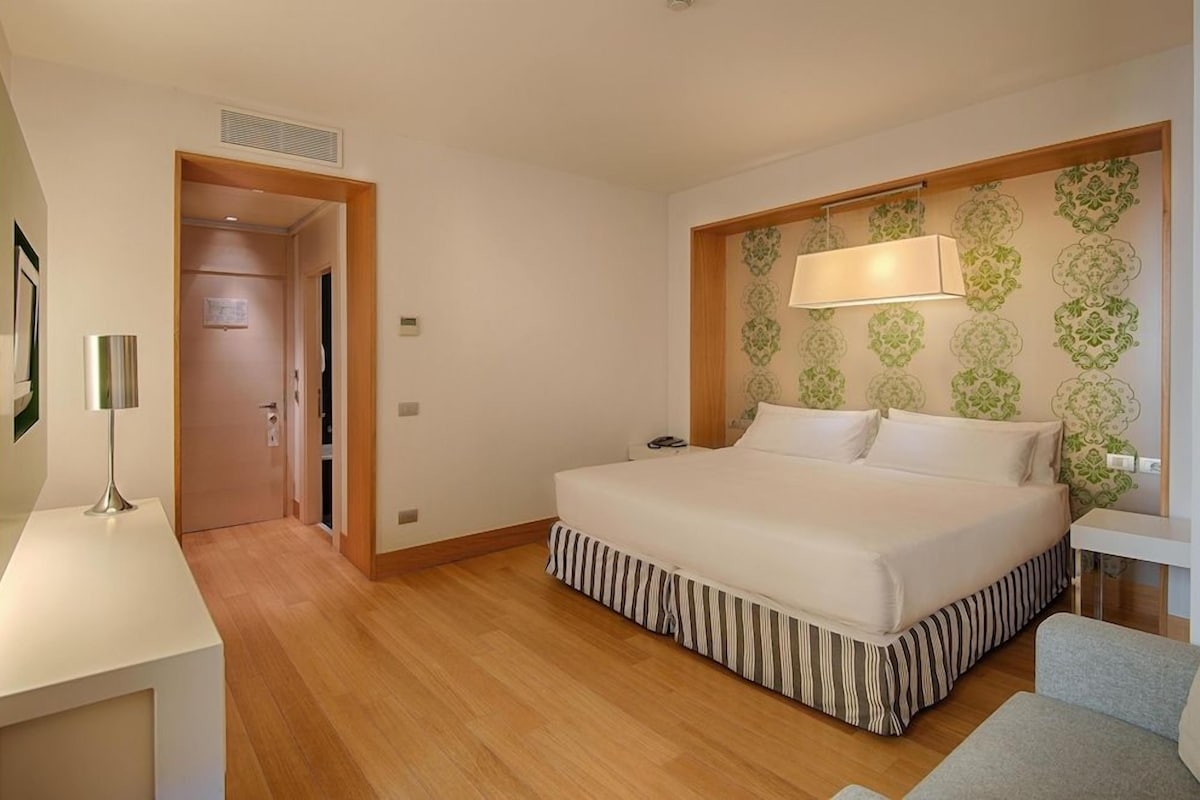 A hotel room with a bed, a desk, a chair, a lamp, and a patterned wallpaper.
