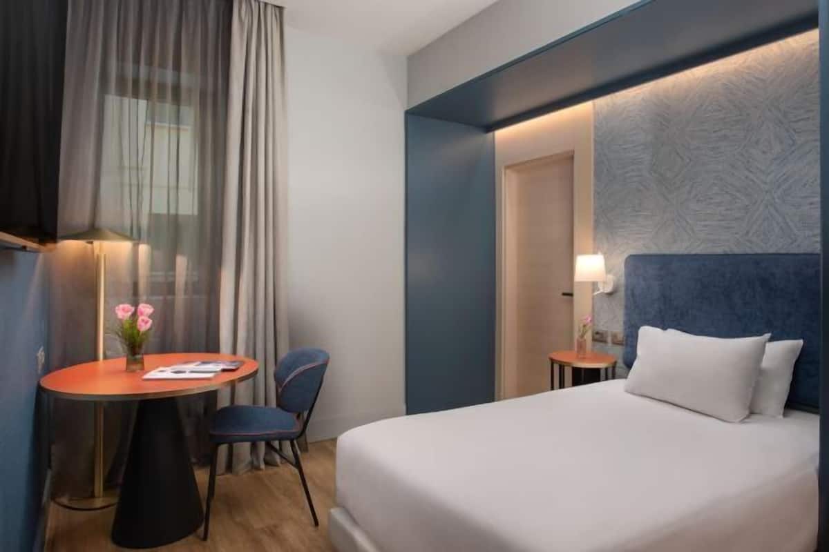 A hotel room with a bed, a desk with a chair, a small table with a chair, and a window with curtains.