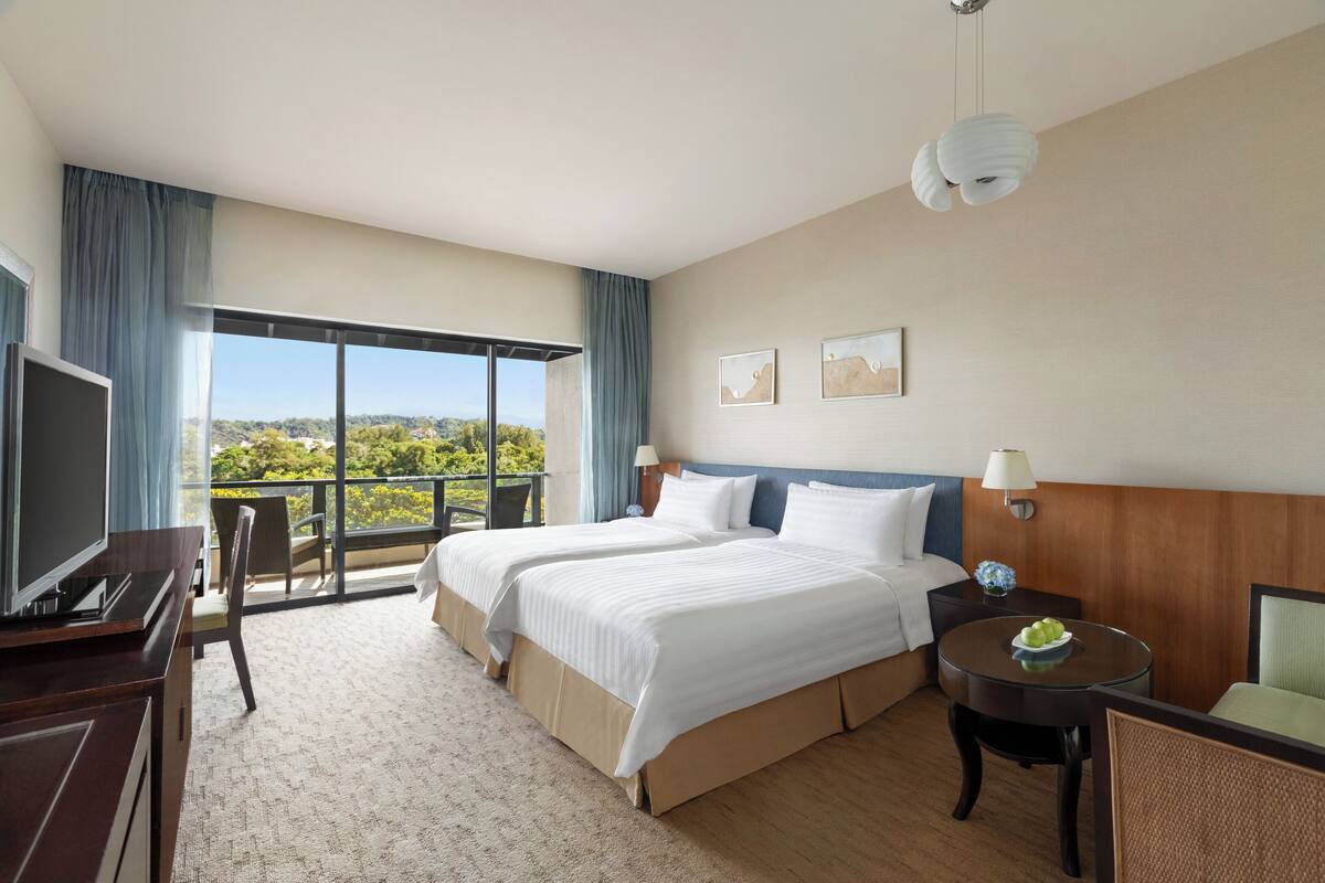 A hotel room with two beds, a TV, a desk with a lamp, and a balcony with a view.