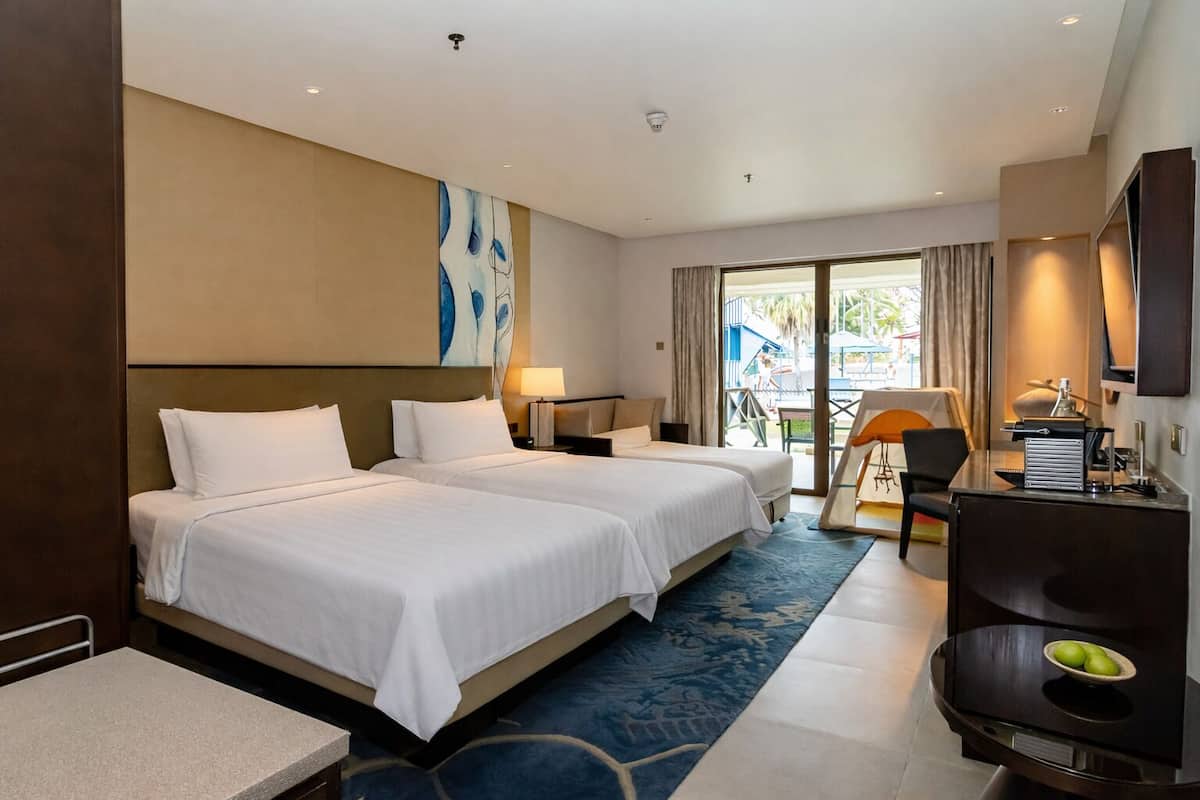 A hotel room with two beds, a desk, a chair, a TV, and a balcony with a view.