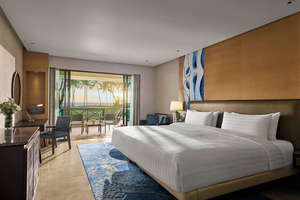 A hotel room with a large bed, a desk with a chair, a desk lamp, a nightstand with a lamp, and a view of the beach.