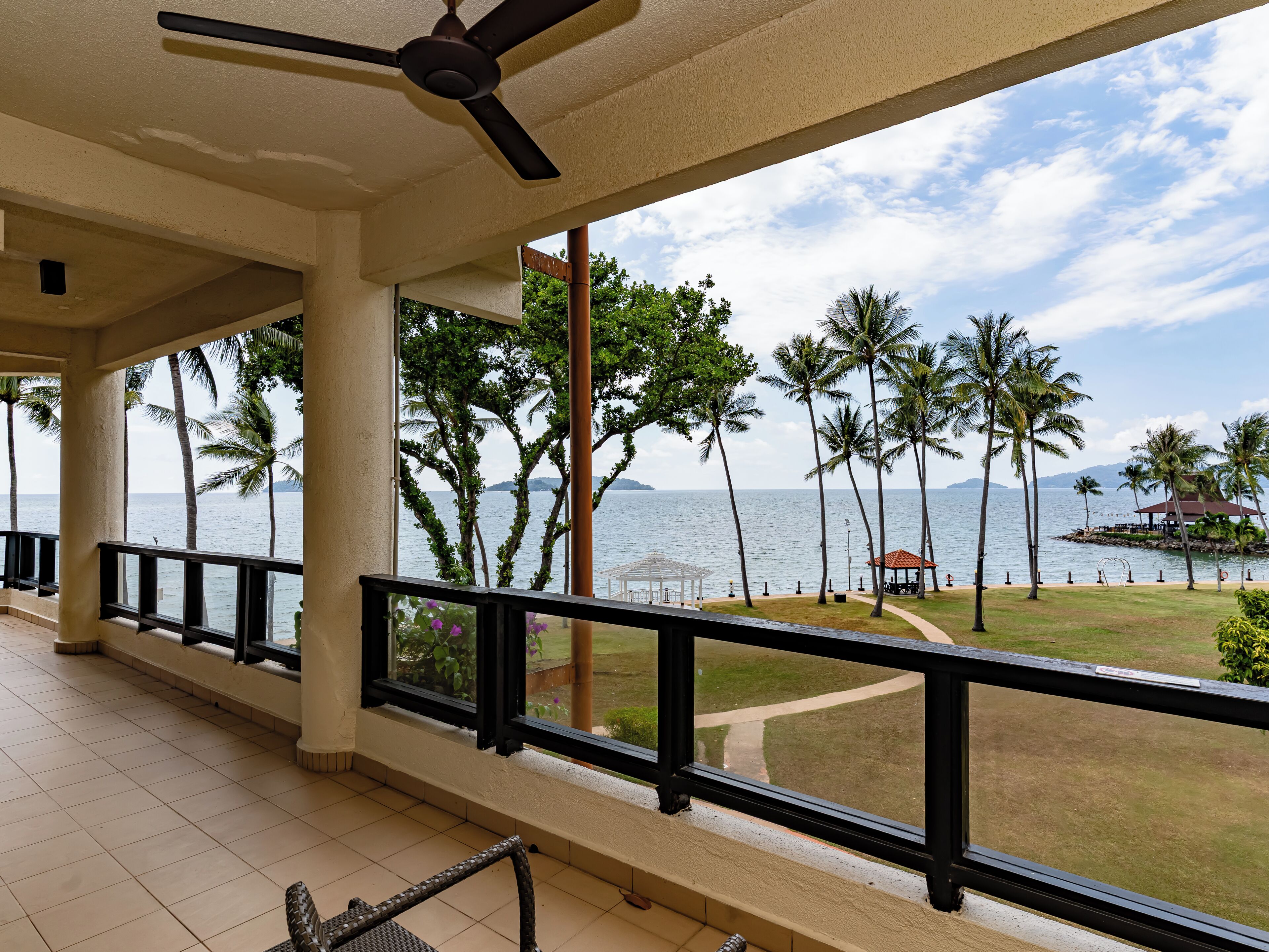 A covered balcony with a ceiling fan, a view of the sea, palm trees, and a gazebo.