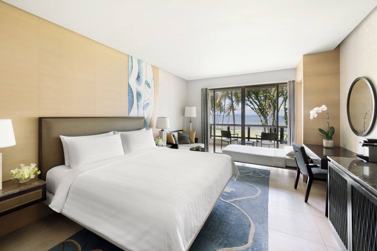 A hotel room with a large bed, a desk, a chair, a desk lamp, a potted plant, a large window with a view of palm trees and the ocean, and a blue patterned rug.