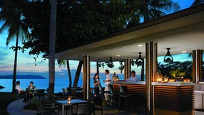 Un espacio de comedor al aire libre con mesas y sillas, un bar y vista al océano al atardecer.