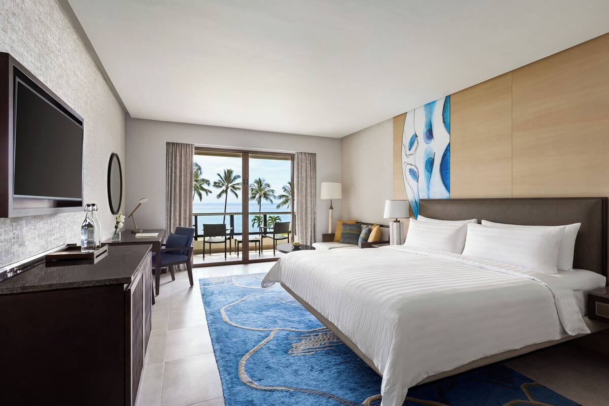 A hotel room with a large bed, a TV, a desk, a chair, a view of the ocean, and a balcony with palm trees.