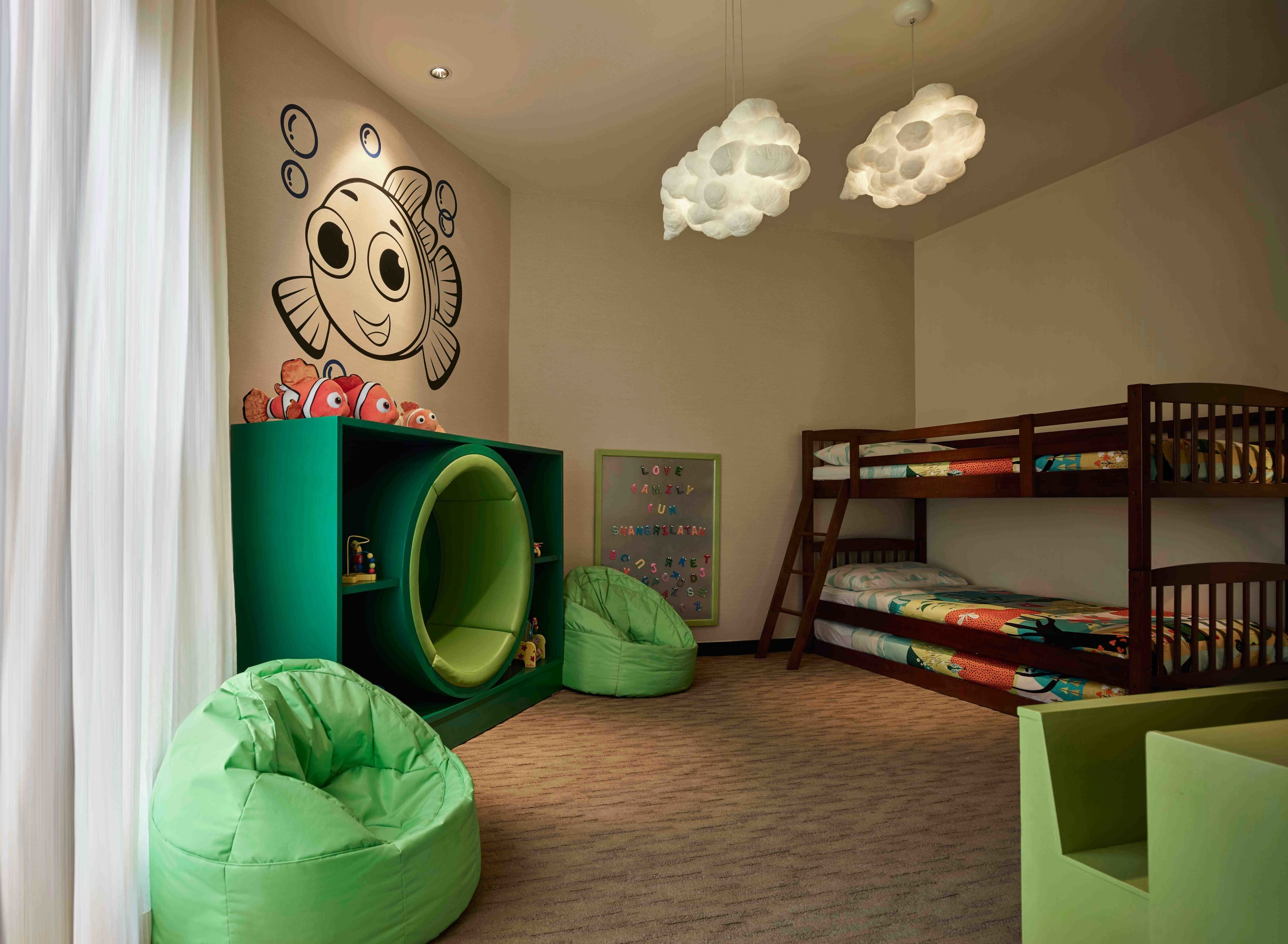 Una habitació infantil amb lliteres, una caseta de joc verda, seients de ploma i una paret amb un personatge de dibuix animat.