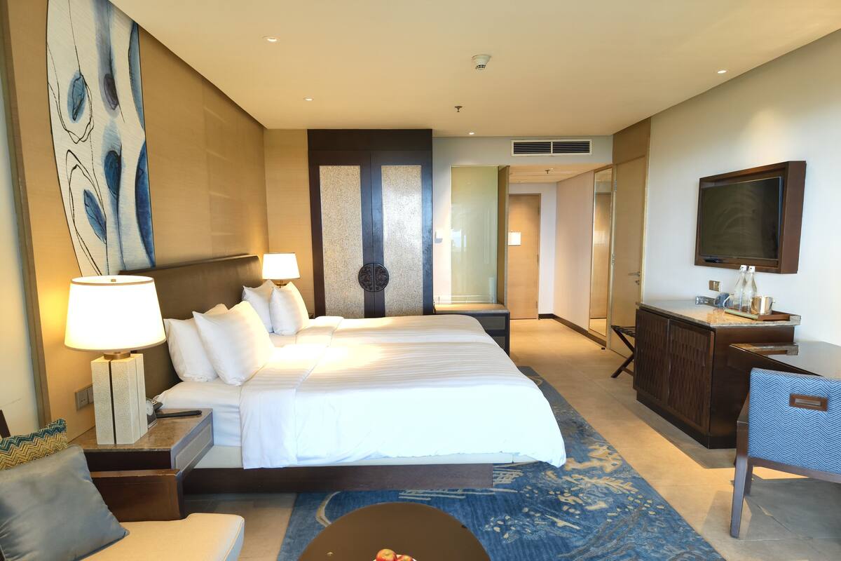 A hotel room with a large bed, a desk, a chair, a TV, and a small table with apples.