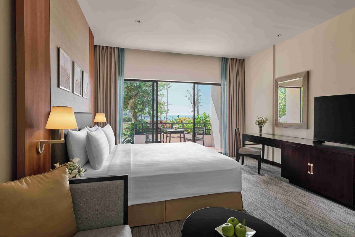 A hotel room with a large bed, a desk with a TV, a sofa, and a balcony with a view.