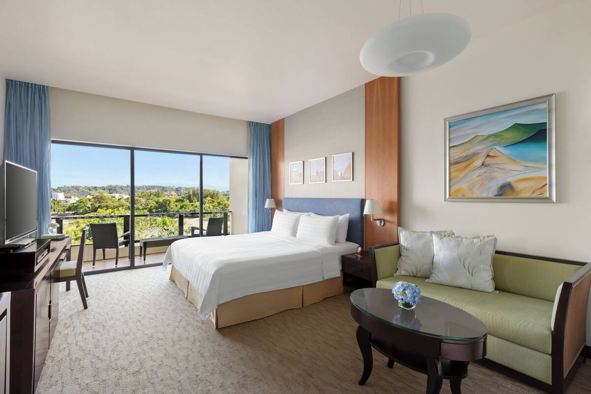 A hotel room with a large bed, a sofa, a desk, and a balcony with a view.