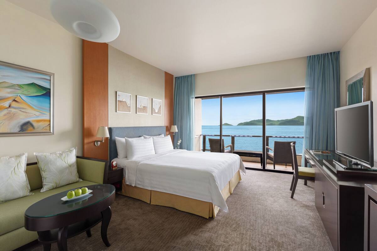 A hotel room with a bed, a sofa, a TV, a balcony with a view of the sea, and a painting on the wall.