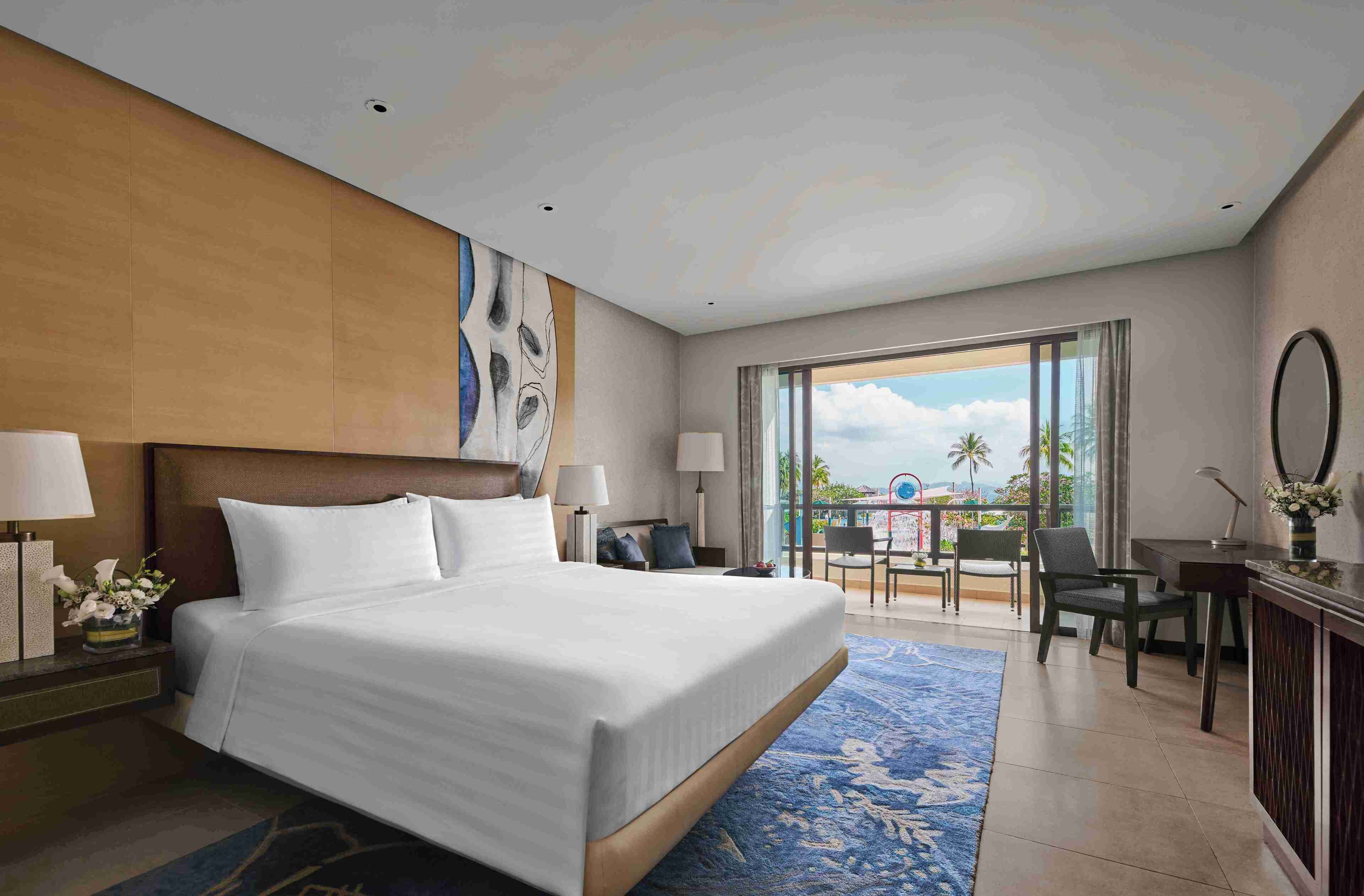 A hotel room with a large bed, a desk, a chair, and a balcony with a view of palm trees.