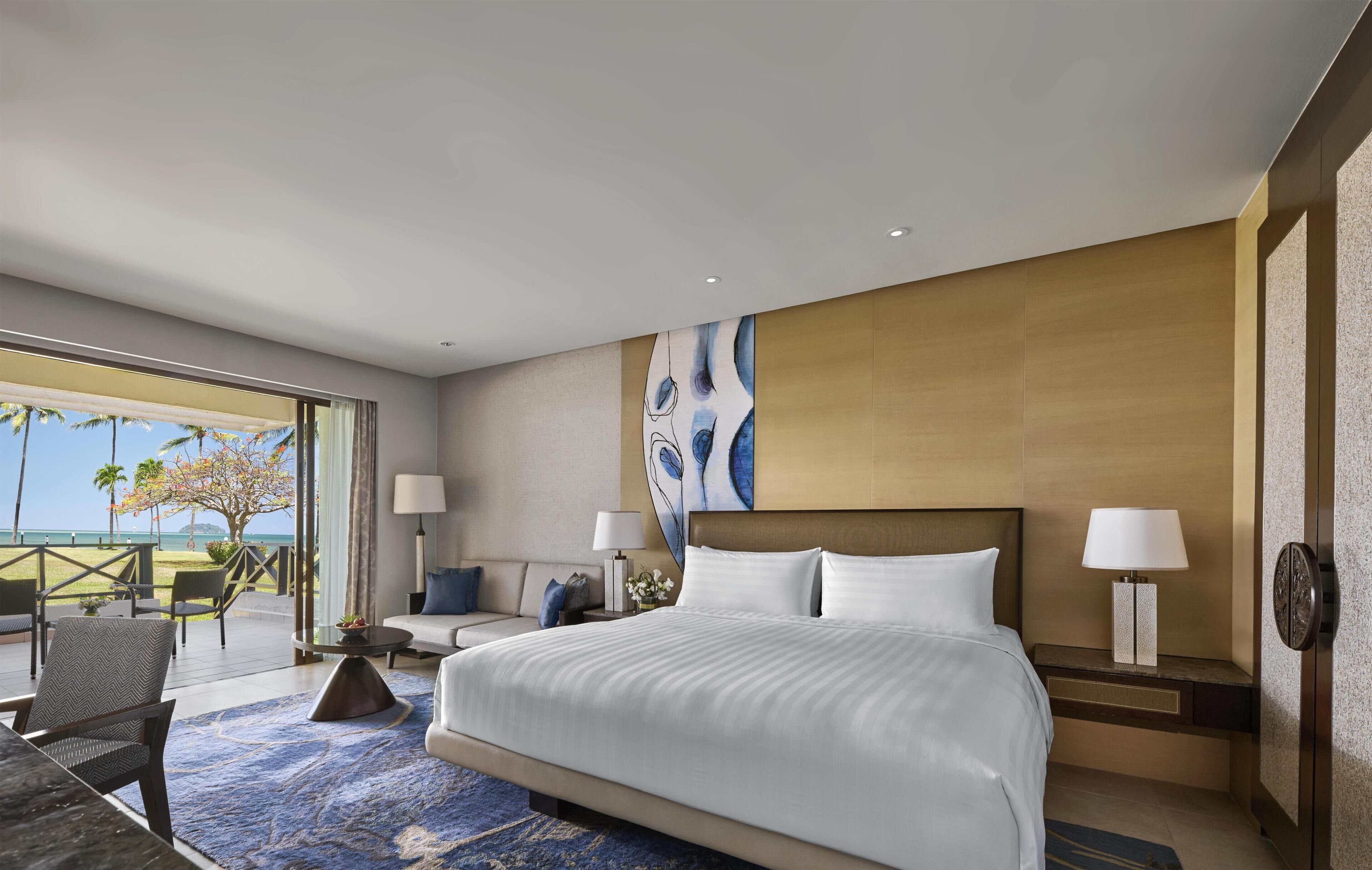A hotel room with a large bed, two bedside tables with lamps, a sofa, a coffee table, and a view of the ocean.