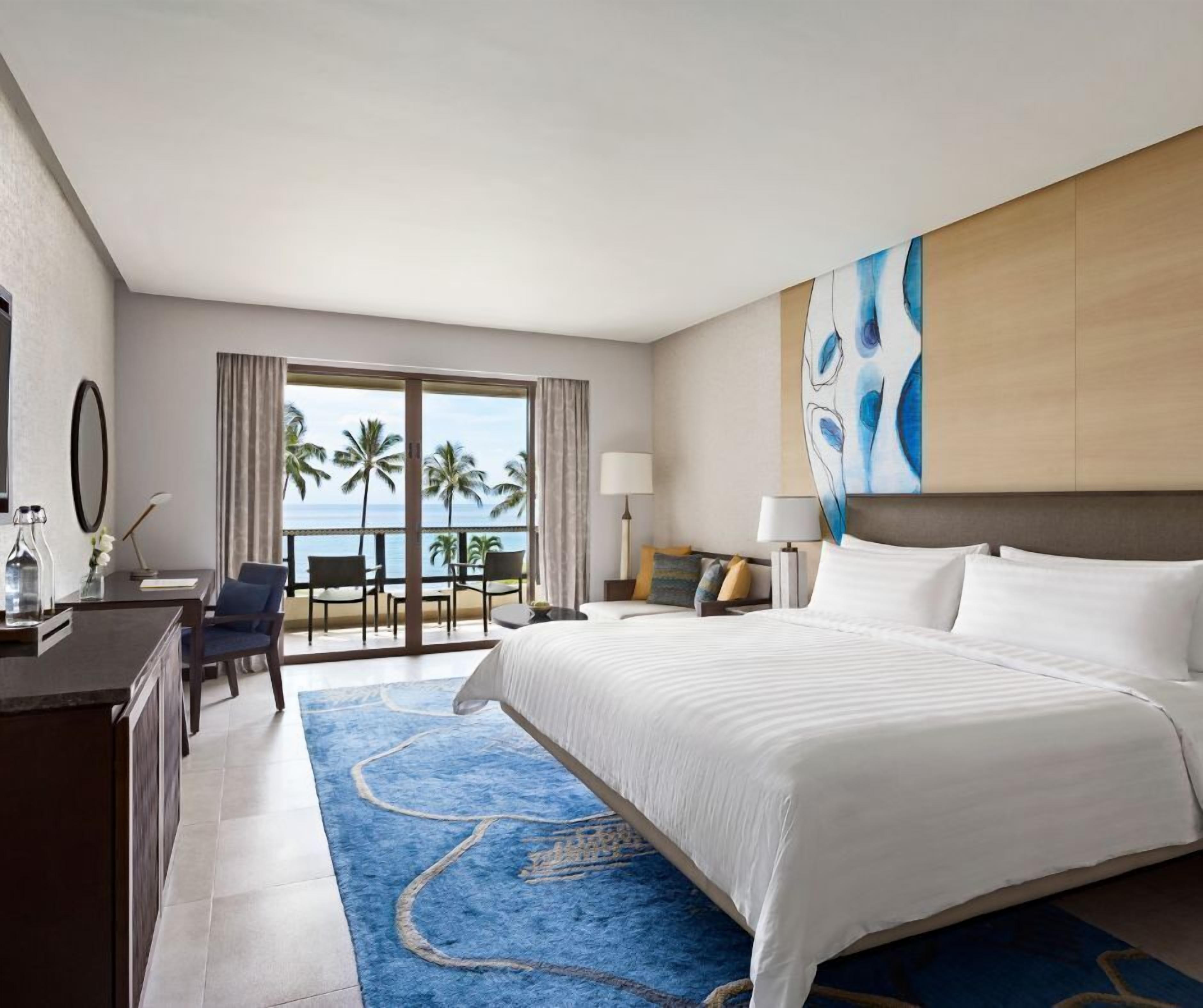 Habitación de hotel con una cama grande, un escritorio con silla, y vistas a las palmeras y al océano a través de las puertas del balcón.
