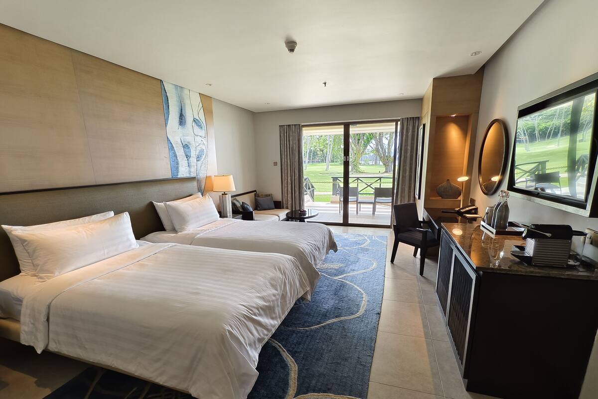 A hotel room with two beds, a desk, a chair, a TV, and a view of the outdoors.
