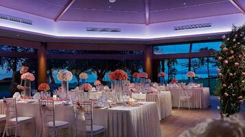 Un salón de recepciones de bodas con mesas redondas, centros de mesa florales y vistas al océano.