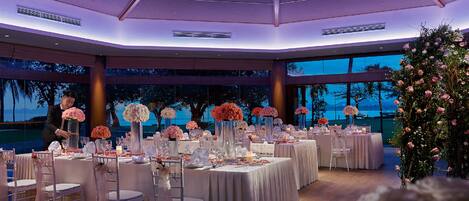 A wedding reception hall with round tables, floral centerpieces, and a view of the ocean.