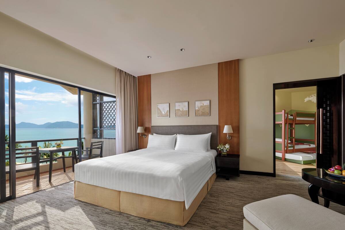A hotel room with a large bed, a balcony with a view of the sea, a desk with a chair, and a small table with fruit.