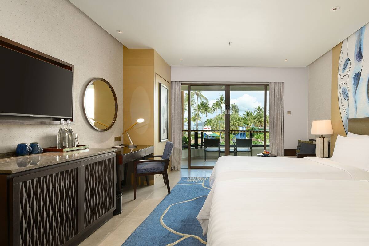 A hotel room with a bed, a desk, a TV, and a view of palm trees.