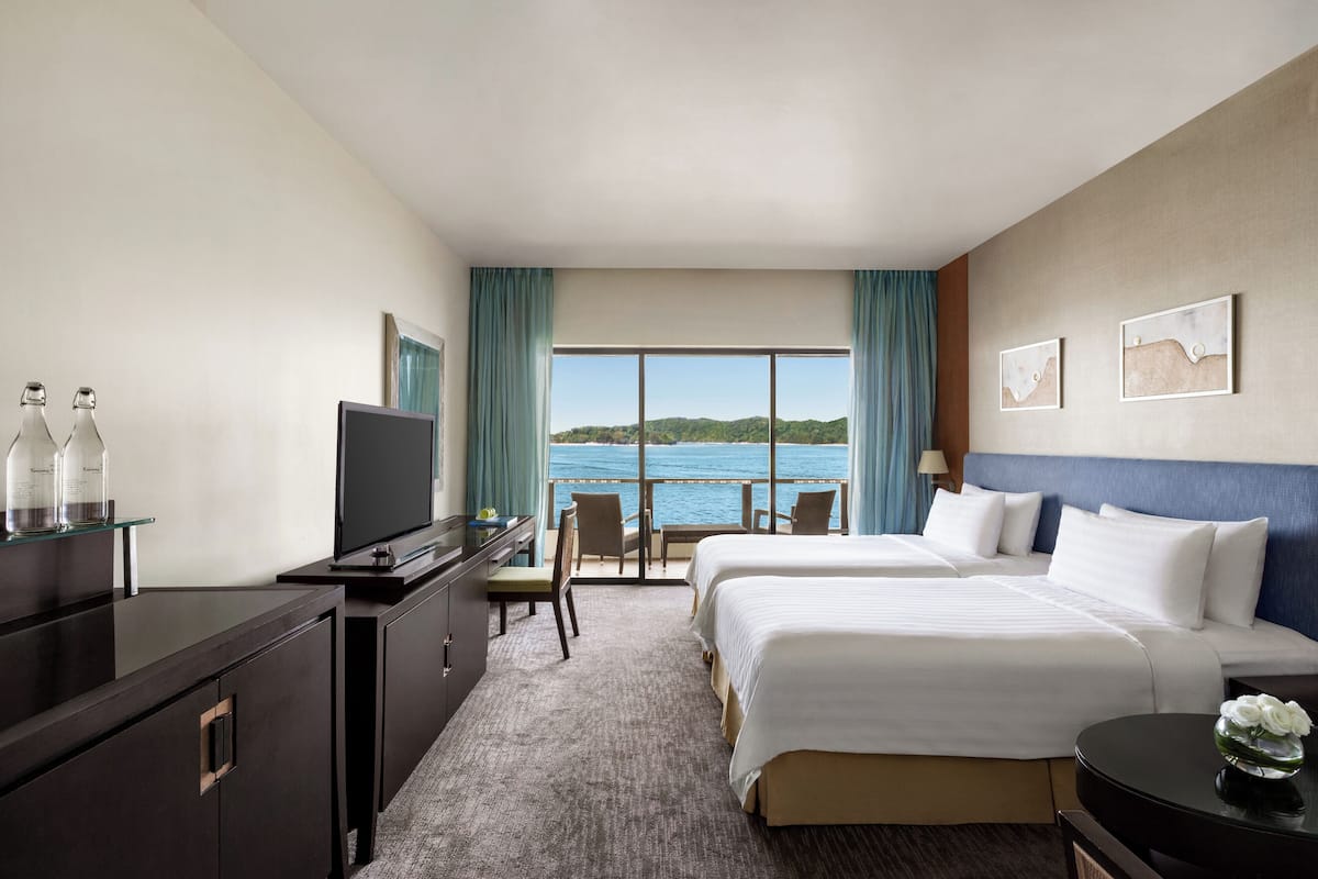 A hotel room with a large bed, a desk with a TV, a chair, and a view of the sea.