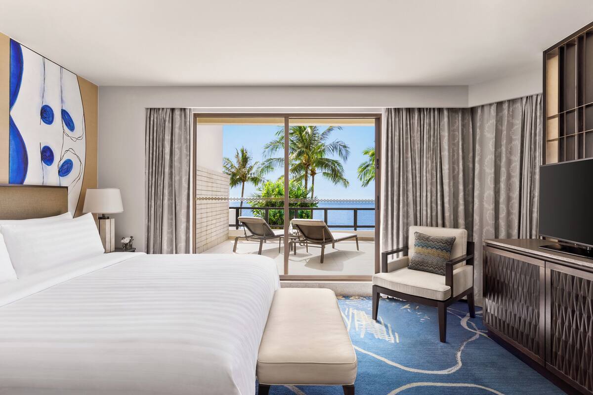 A hotel room with a bed, a TV, a chair, a bench, and a balcony with a view of palm trees and the ocean.