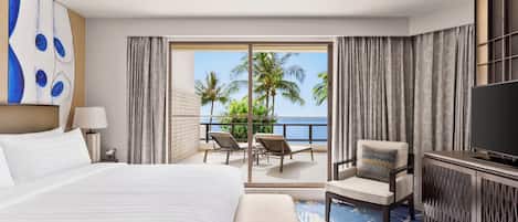 A hotel room with a large bed, a TV, a seating area with a view of palm trees and the sea, and a balcony with curtains.