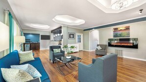 A modern living room with a blue sofa, a grey armchair, and a glass coffee table. There is a fireplace on the wall and a kitchen area in the background. - Best Western Post Oak Inn (Burnet)