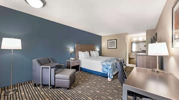 A hotel room with a bed, a desk, a chair, a desk lamp, a floor lamp, and a small kitchenette.